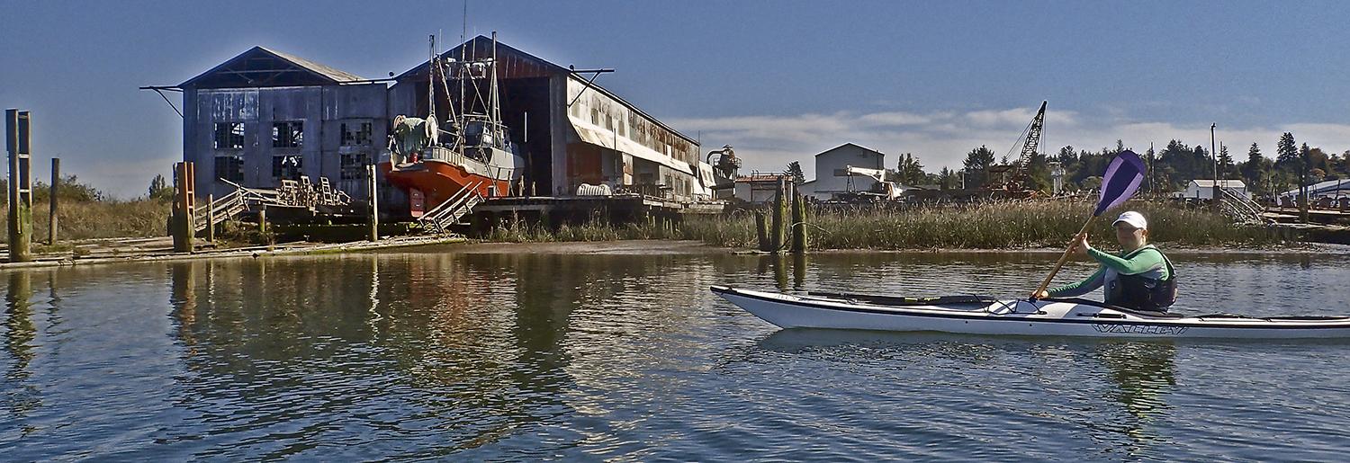 woman kayaks past a fishingboat in drydock at an old, rusting building