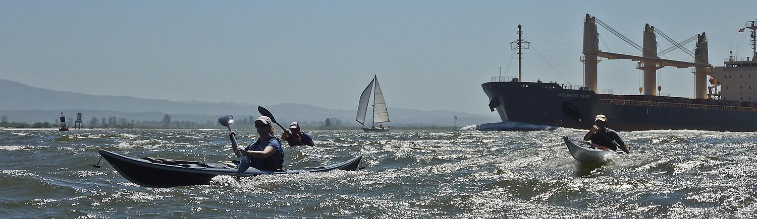 three kayakers paddle through choppy water, with a barge and sailboat in the distance