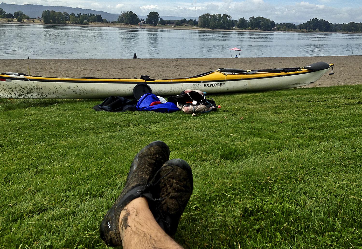 the author's ankles and feet, crossed, resting on the grassy shore looking at the kayak, sandy beach, and river