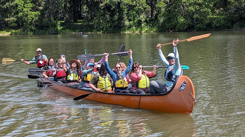 a group of 14 people in a big canoe - some hold their paddles up in greeting